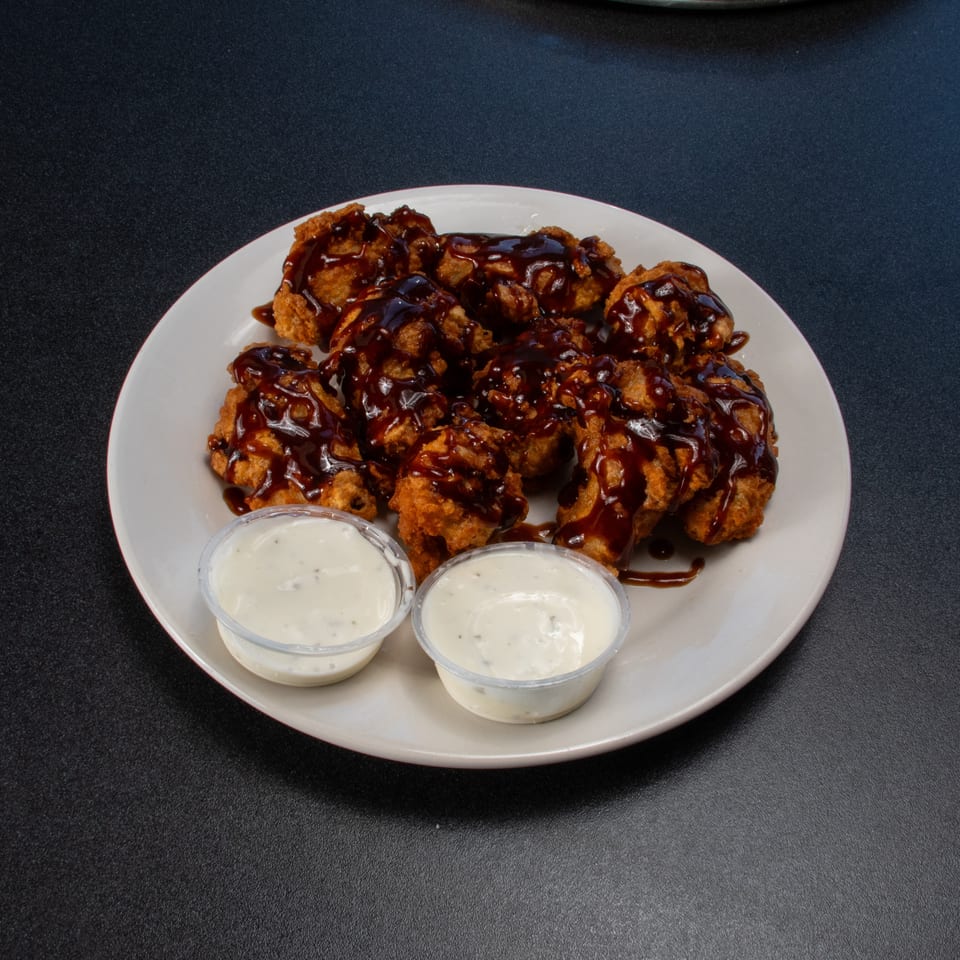 Plate of crispy fried bites drizzled with dark sauce, served with two cups of creamy white dipping sauce.