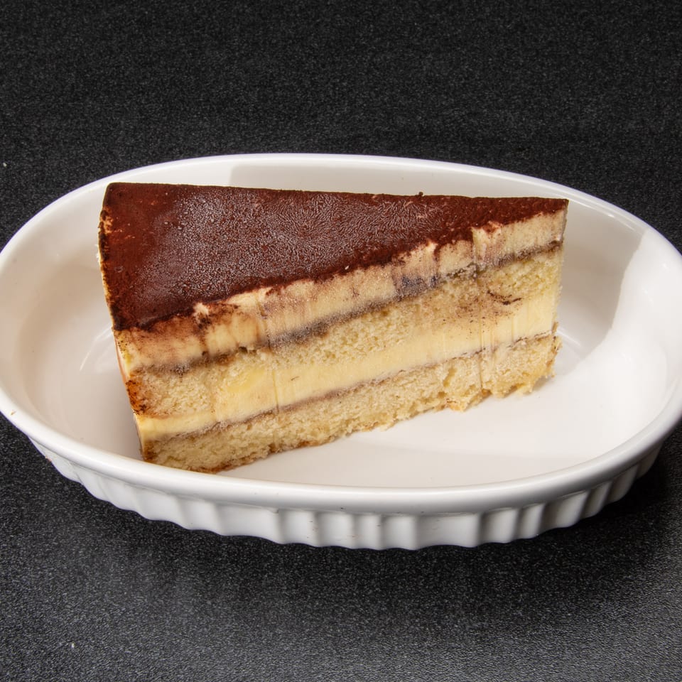 Slice of layered vanilla cake topped with cocoa powder, served on a white oval plate.