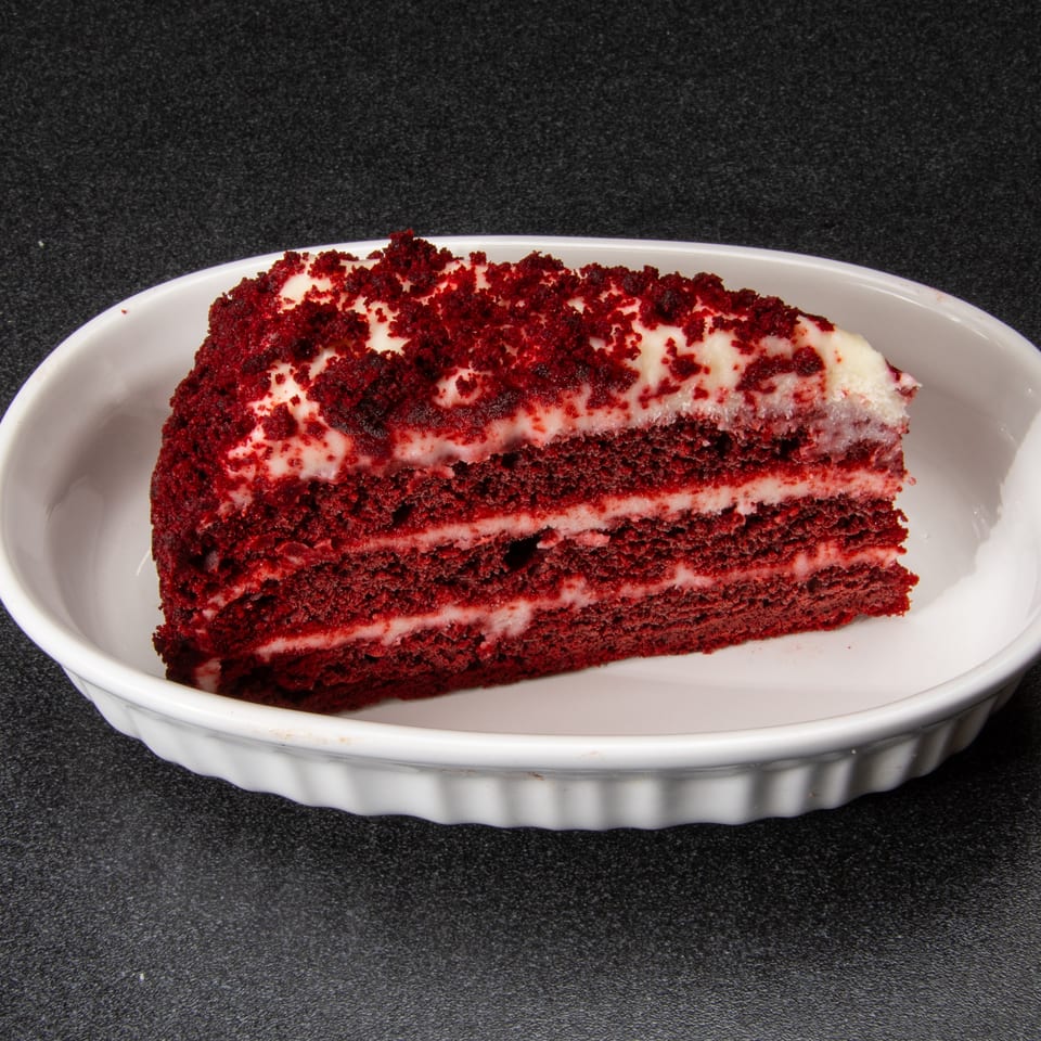 Slice of red velvet cake with white frosting and red crumbs on a white serving dish.