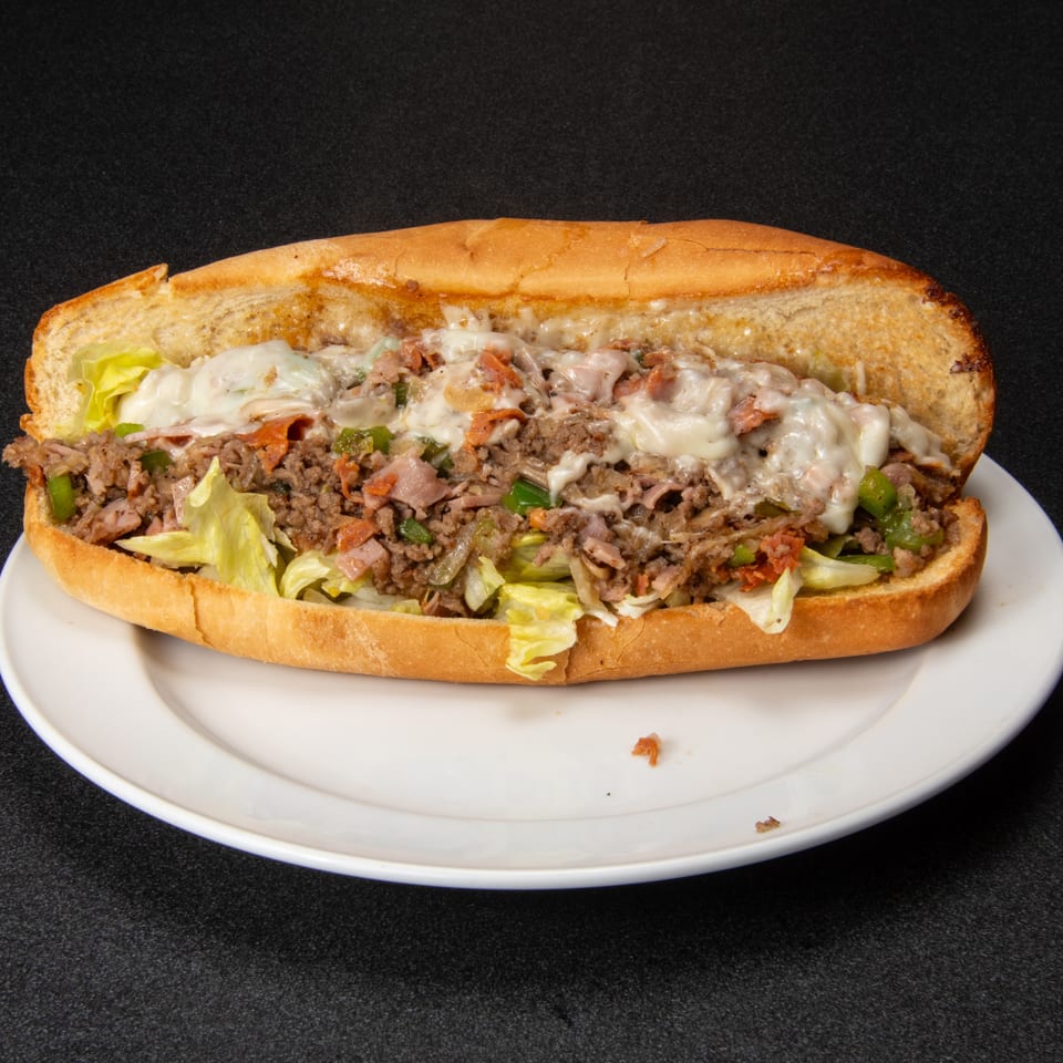 Loaded steak and cheese hoagie with melted cheese, bacon, lettuce, onions, and green peppers on a white plate.